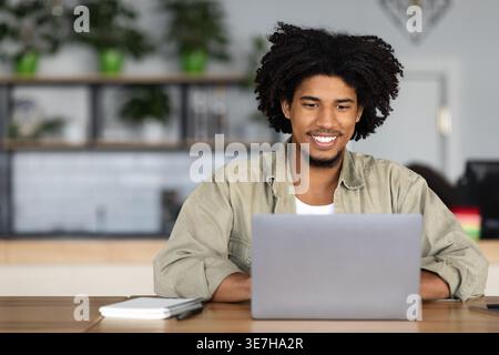 Smiling Black male with afro using laptop sitting at table in cafe Stock Photo
