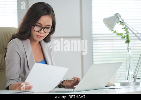 Asian business woman wearing eyeglasses reading documents while working on laptop sitting at table in office Stock Photo