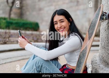 Venezuelan female skater with long black hair holding smartphone sitting on street during daytime Stock Photo