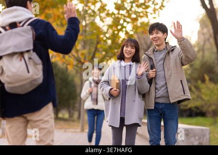 Cheerful Chinese male and female college students waving hands greeting male classmate walking on campus in afternoon Stock Photo