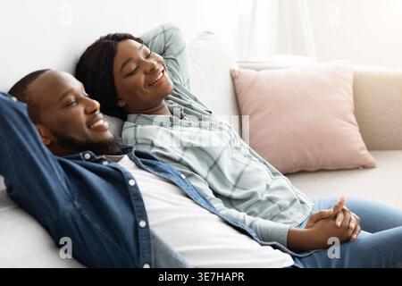Black husband and wife holding hands while sitting with closed eyes on sofa in living room at home Stock Photo