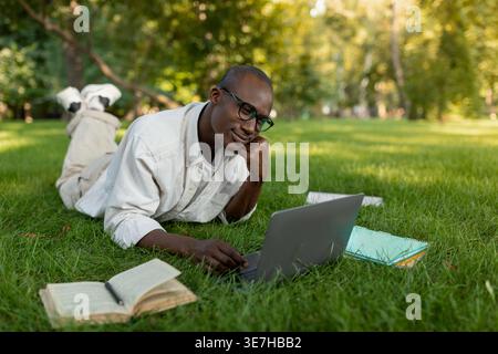 Black male student wearing eyeglasses leaning face on hand using laptop while lying on grass near book and notepads in campus park during daytime Stock Photo