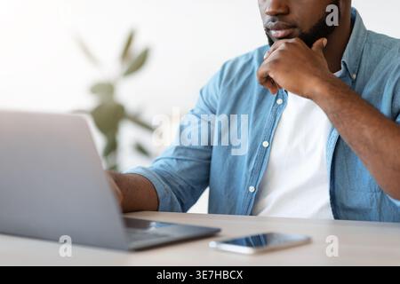 Cropped image of bearded Black male freelancer leaning face on hand working on laptop while sitting at table in home office during daytime Stock Photo