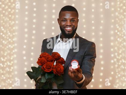 Smiling Black man holding red roses and box with ring standing against festive background Stock Photo