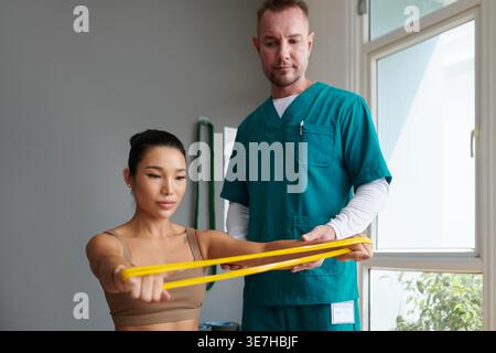 Vietnamese female patient doing arm stretching exercise using resistance band with caucasian male physiotherapist assisting her in clinic Stock Photo