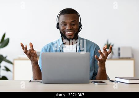 Smiling bearded Black male freelancer wearing headset having videocall on laptop gesturing hands sitting at table with smartphone in home office Stock Photo