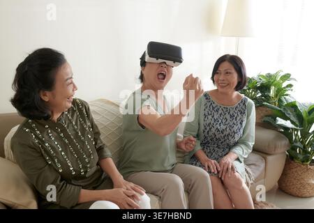Asian senior woman using VR glasses sitting on sofa beside senior friends looking at her in living room at home Stock Photo