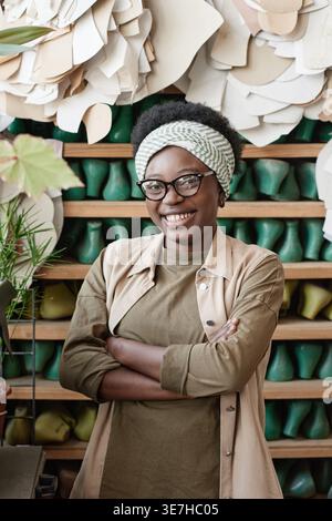 Black female tailor wearing eyeglasses while standing with crossed arms in front of shelf in workshop Stock Photo