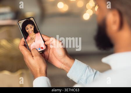 Back over-the-shoulder view of Black man having video call on smartphone with his surprised girlfriend indoors Stock Photo