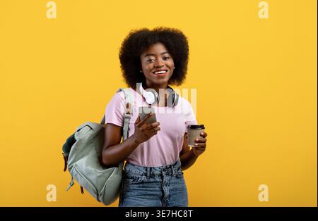 Afro-haired Black female student with headphones around neck and backpack holding smLifestylephone and coffee cup standing against yellow background in s... Stock Photo