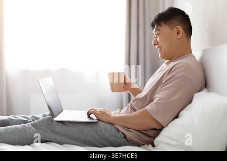 Relaxed happy cheerful middle aged handsome chinese man wearing pajamas chilling on bed, using laptop, holding mug, drinking coffee, enjoying weekend Stock Photo
