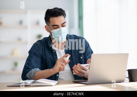 Middle Eastern man wearing protective face mask using disinfectant for hands sitting at table with laptop at home Stock Photo