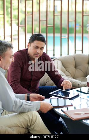 Asian businessman using tablet talking with white male colleague sitting on couch in office lounge Stock Photo