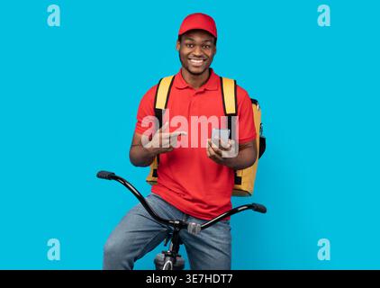 Smiling Black woman wearing cap pointing finger on smartphone sitting on bicycle against blue background in studio Stock Photo