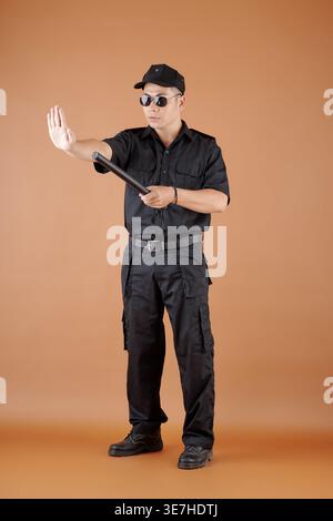 Asian male police officer in black uniform, cap and sunglasses holding baton making stop gesture standing against brown background in studio Stock Photo