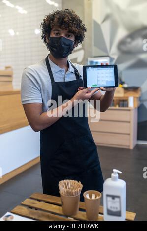 Asian male waiter with curly hair wearing protective mask showing tablet while standing in coffee shop Stock Photo