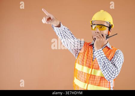 Asian male civil engineer in vest and hardhat pointing away talking on walkie talkie standing against brown background in studio Stock Photo