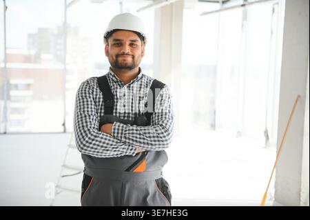 Indian male builder wearing uniform and hard hat standing with crossed arms on construction site Stock Photo