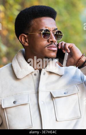 Hispanic man wearing sunglasses having phone call and looking away standing outdoors during daytime Stock Photo