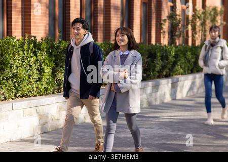 Smiling male and female college students wearing jackets walking together near bushes on campus on sunny day Stock Photo