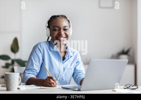 Black business woman with braids wearing headphones using laptop taking notes while sitting at table in home office Stock Photo