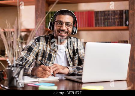 Indian male student in headphones wearing eyeglasses writing down notes learning online using laptop sitting at table in library Stock Photo