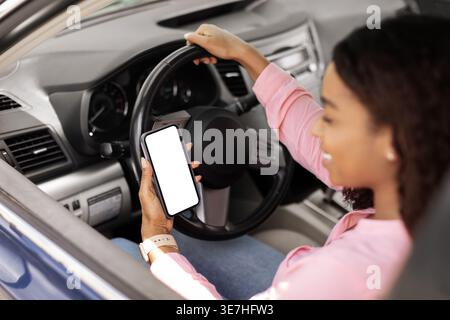 Smiling Black woman wearing pink shirt using smartphone while sitting on driver seat holding steering wheel in car Stock Photo