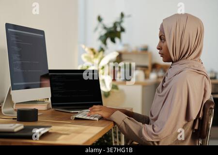 Black female programmer in hijab working codes on computer while sitting at table at home office Stock Photo