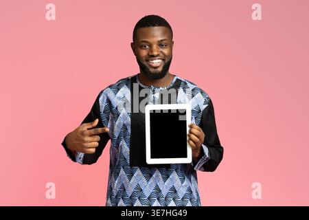 Smiling black guy pointing at digital tablet with empty screen, wearing traditional african costume. Positive african american young man showing newes Stock Photo
