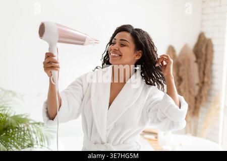 Black woman in bathrobe blow drying hair standing in bathroom at home Stock Photo