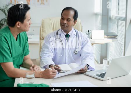 Asian male doctors in scrub suit and white lab coat with stethoscope around neck having conversation holding paper sitting at table in hospital office Stock Photo