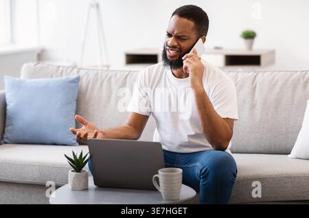 Black man with laptop talking on smartphone while sitting on sofa in living room at home Stock Photo