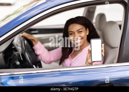Smiling Black woman wearing pink shirt showing smartphone while sitting on driver seat holding steering wheel in car Stock Photo