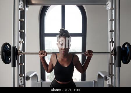 Black woman in fitness wear wearing earbuds listening to music doing squats with barbell on her shoulders in smith machine at gym Stock Photo
