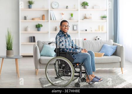 Smiling Black disabled man wearing eyeglasses sitting on wheelchair in living room at home Stock Photo