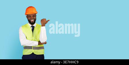 Black male engineer wearing hard hat and safety vest pointing thumb sideways standing with crossed arm against blue background in studio Stock Photo