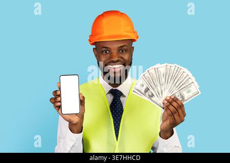 Black male engineer wearing hard hat and safety vest showing money and smartphone with blank screen while standing against blue background in studio Stock Photo