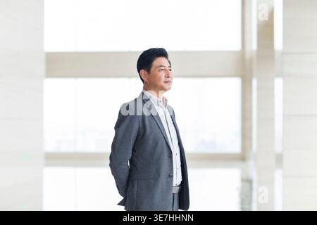 Serious Chinese businessman wearing gray suit looking away while standing with hands behind back against window in office Stock Photo