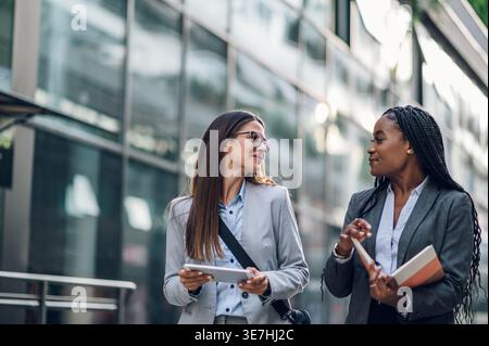 Smiling Black businesswoman with braids holding book talking to caucasian female colleague with tablet walking outside office building during daytime Stock Photo