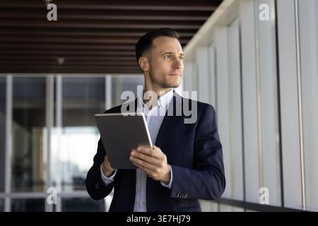 Serious businessman holding digital tablet, looking out office window thoughtfully Stock Photo
