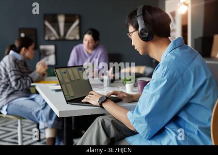 Side view of Asian man with headphones working on laptop screen while sitting at table with colleagues in office Stock Photo