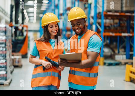 Black male worker wearing hard hat and safety vest holding clipboard and managing inventory with caucasian female coworker while standing in warehouse Stock Photo