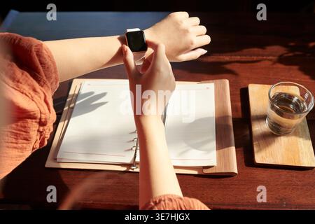 Hands of Asian woman checking notifications on smartwatch over the table with notebook and glass of water indoors Stock Photo