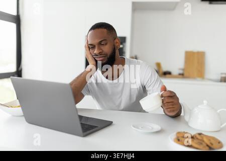 Bored Black man leaning face on hand holding coffee cup sitting at counter with laptop at home Stock Photo