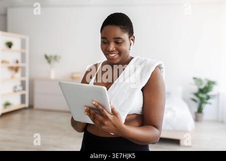 Curvy Black woman in wireless earphones and towel on shoulder using tablet while standing in bedroom at home Stock Photo