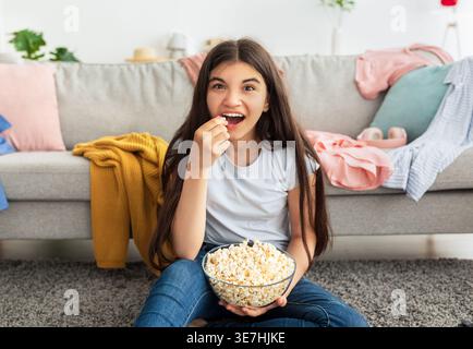 Happy mixed-race female teenager eating popcorn sitting on floor carpet in living room at home Stock Photo