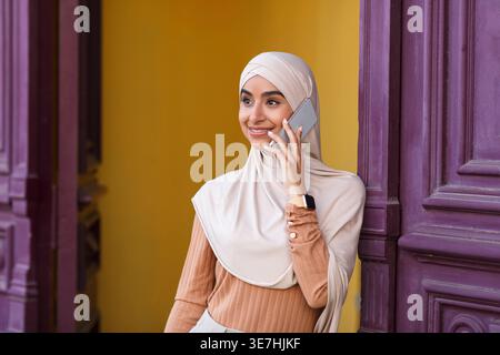 Smiling Indian Muslim woman wearing hijab looking away talking on smartphone standing near front door at home during daytime Stock Photo