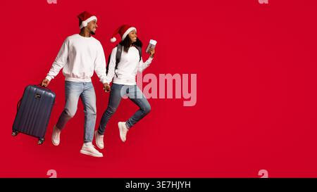 Smiling Black couple with backpack and luggage wearing Santa hats looking away holding passport with tickets running against red background in studio Stock Photo