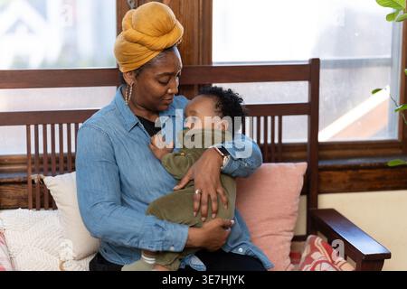 Black mother wearing yellow head wrap and long sleeve denim shirt carrying and playing with her baby boy with pacifier while sitting on wooden sofa... Stock Photo