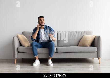 Middle Eastern man looking away having phone conversation sitting on sofa in living room at home Stock Photo
